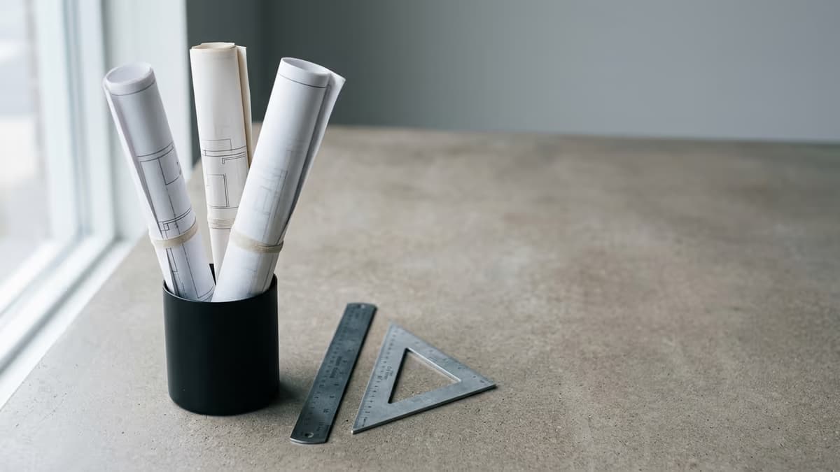 Rolled architectural drawings standing beside a metal scale ruler and drafting triangle in a design studio, representing a licensed architecture and engineering firm