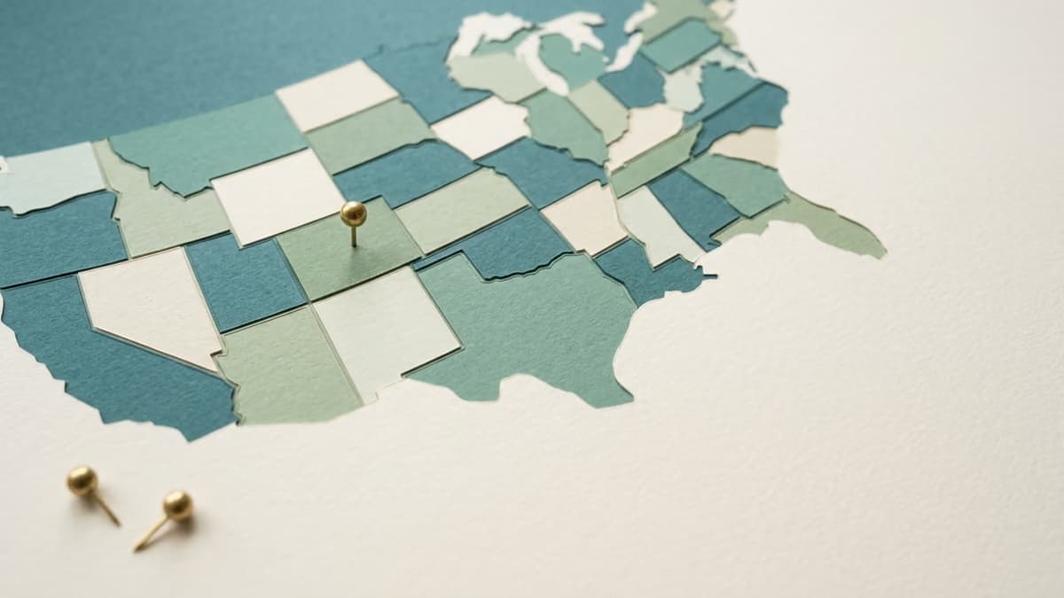 Layered paper map of the United States with a single brass pin pressed into one state and two loose pins resting beside the map