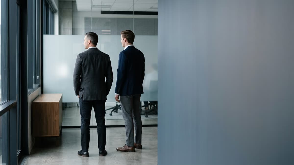 Two business professionals standing side by side in a modern office, one a step ahead of the other