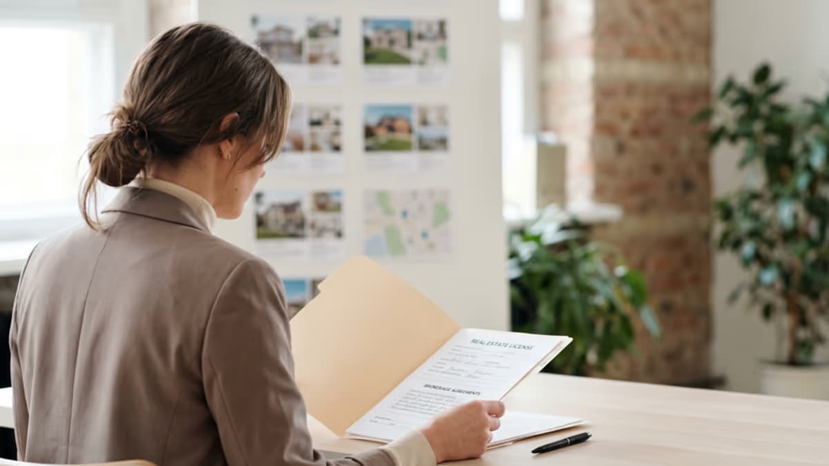 An over-the-shoulder view of a real estate professional reviewing a license folder and brokerage documents in a modern real estate office in soft daylight, modern editorial composition with negative space on the right