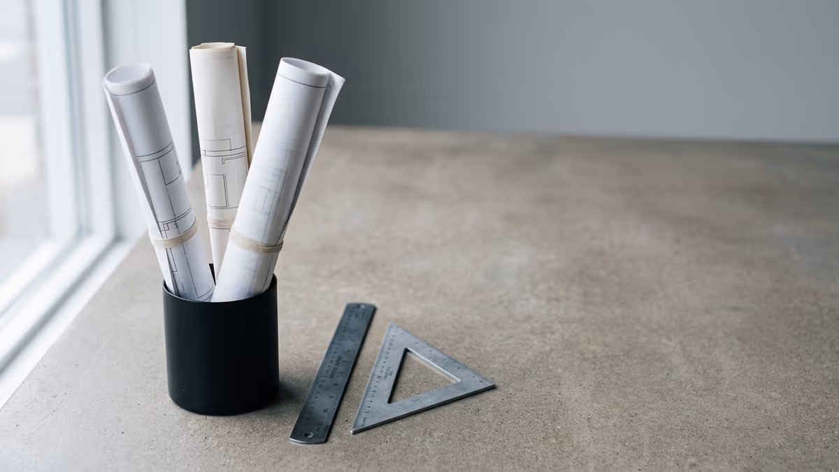Rolled architectural drawings standing beside a metal scale ruler and drafting triangle in a design studio, representing a licensed architecture and engineering firm