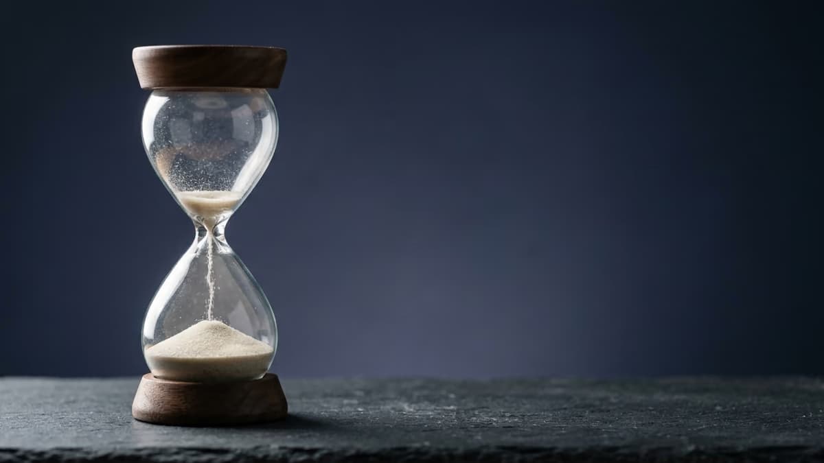 A glass hourglass on a dark slate surface with almost all of its sand run into the lower bulb, against a deep indigo backdrop