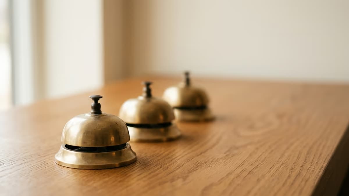 Three small brass service bells arranged in a row on a warm wooden surface, with only the nearest one in sharp focus while the others soften into the background under gentle window light, suggesting that the answer depends on which desk you approach.
