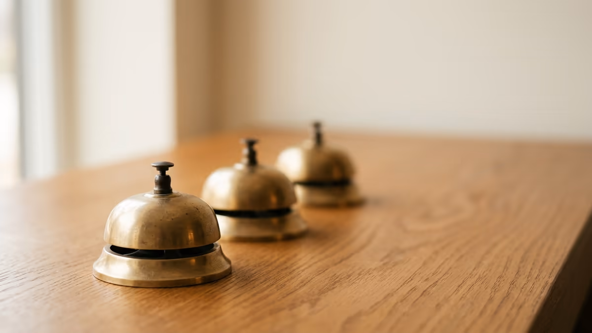 Three small brass service bells arranged in a row on a warm wooden surface, with only the nearest one in sharp focus while the others soften into the background under gentle window light, suggesting that the answer depends on which desk you approach.