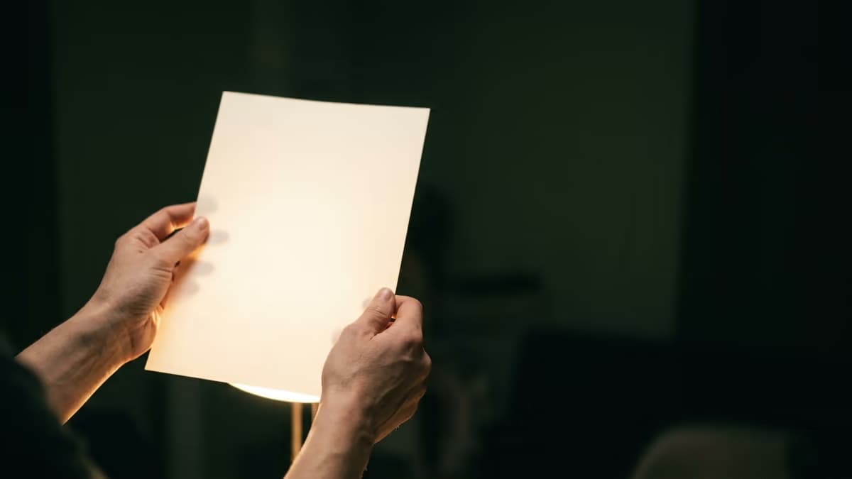 Hands holding a single sheet of paper up to a lamp in a darkened room, the page glowing but revealing nothing.