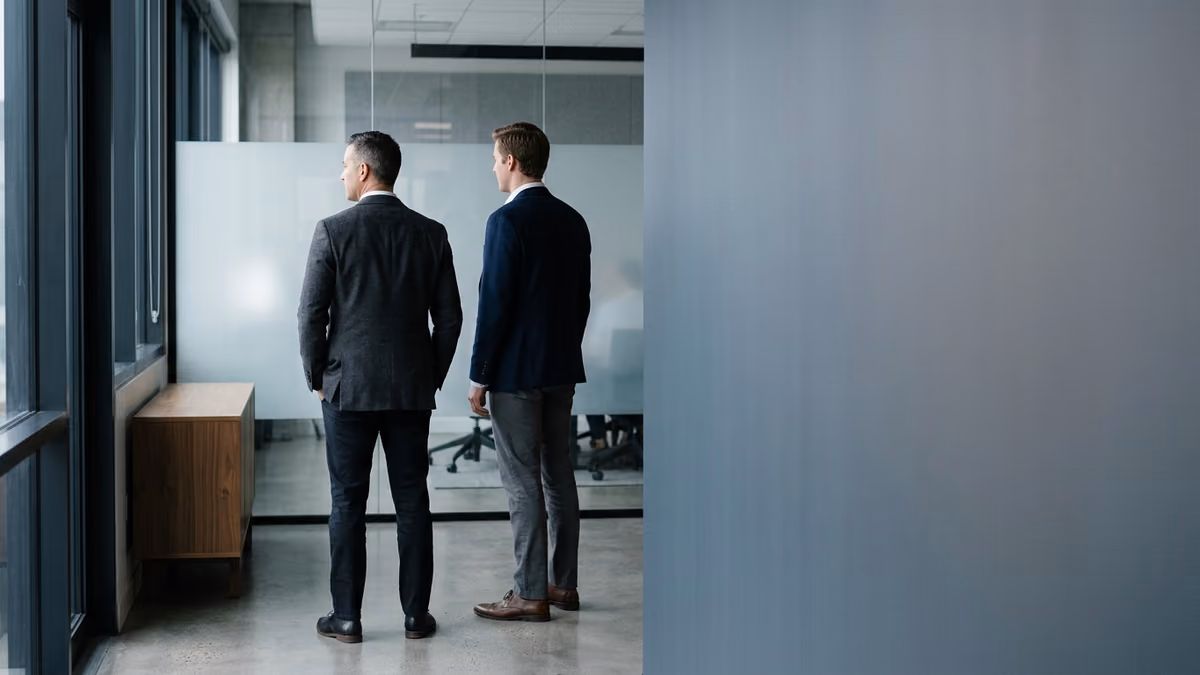 Two business professionals standing side by side in a modern office, one a step ahead of the other