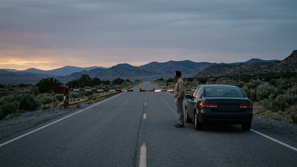 A lone driver standing beside a parked car at a lowered red and white barrier on an empty Nevada desert highway at dusk
