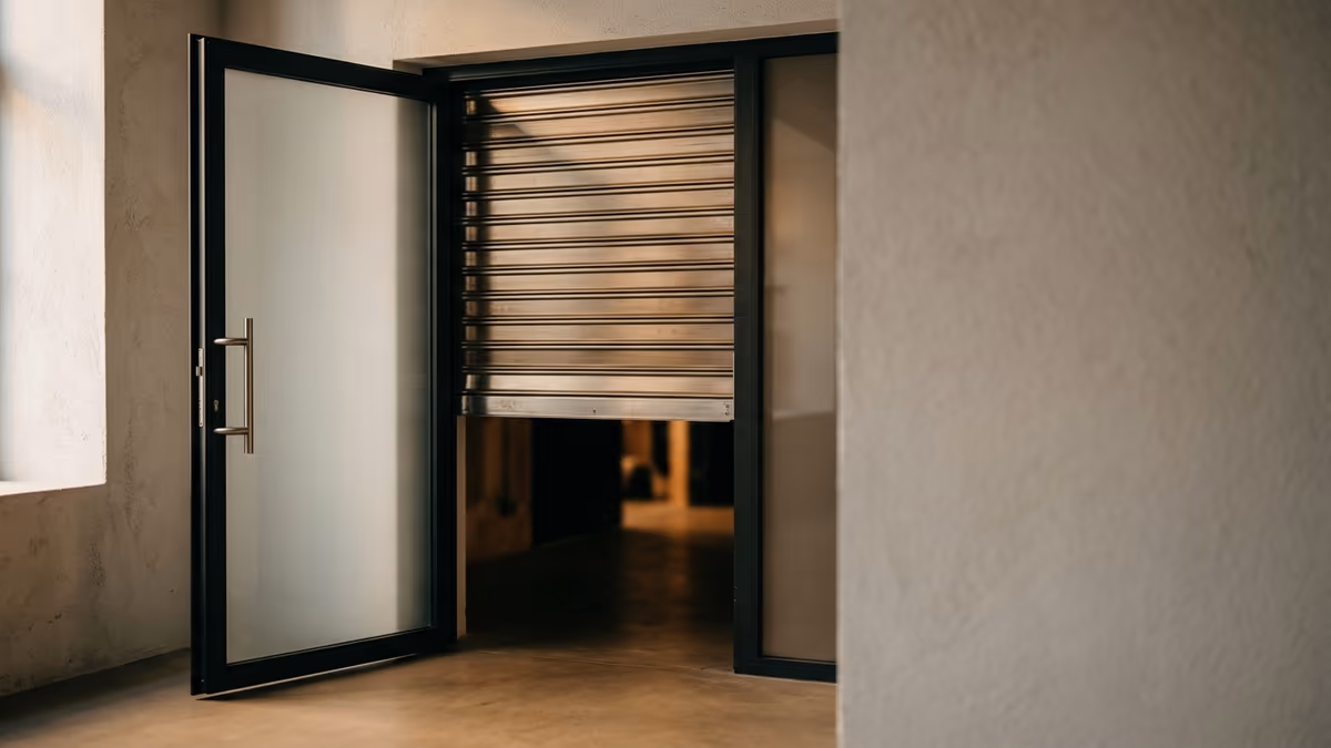 A half-lowered metal security shutter positioned just behind an open glass doorway, lit by warm directional light, symbolizing a second gate that can close after an account is approved.