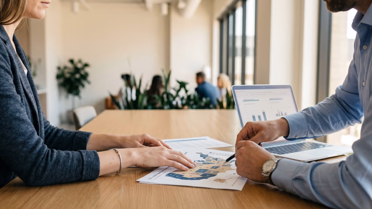 Two business professionals reviewing a multi-state tax map at a modern office conference table, side angle, natural window light, with an open laptop and a printed state-by-state breakdown between them, editorial composition