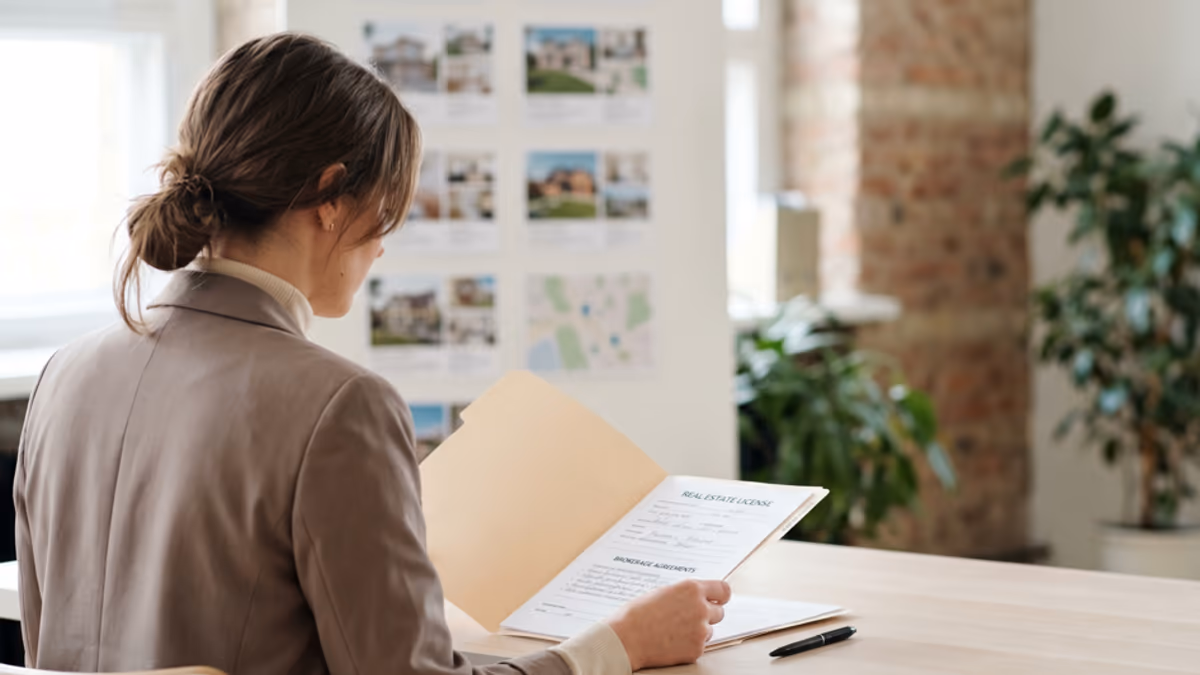 An over-the-shoulder view of a real estate professional reviewing a license folder and brokerage documents in a modern real estate office in soft daylight, modern editorial composition with negative space on the right