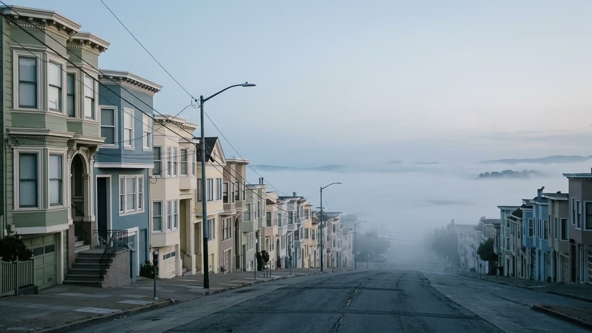 A steep San Francisco residential street lined with pastel row houses descending toward a fog-covered bay at dawn
