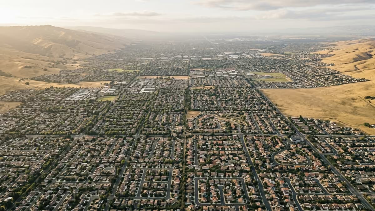 An aerial view of a sunlit valley where neighborhood street grids change direction from one area to the next, meeting dry golden hills at the edge