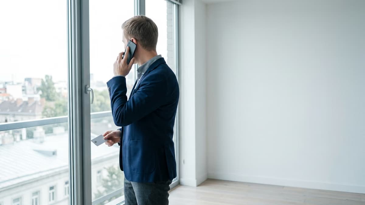 A business professional standing at a bright window, phone to one ear and a plain unbranded payment card in the other hand, calling the card issuer